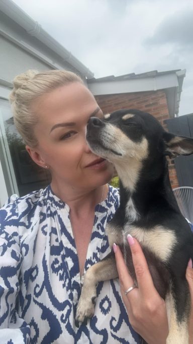 Woman in a patterned shirt kissing a small black and tan dog outdoors.